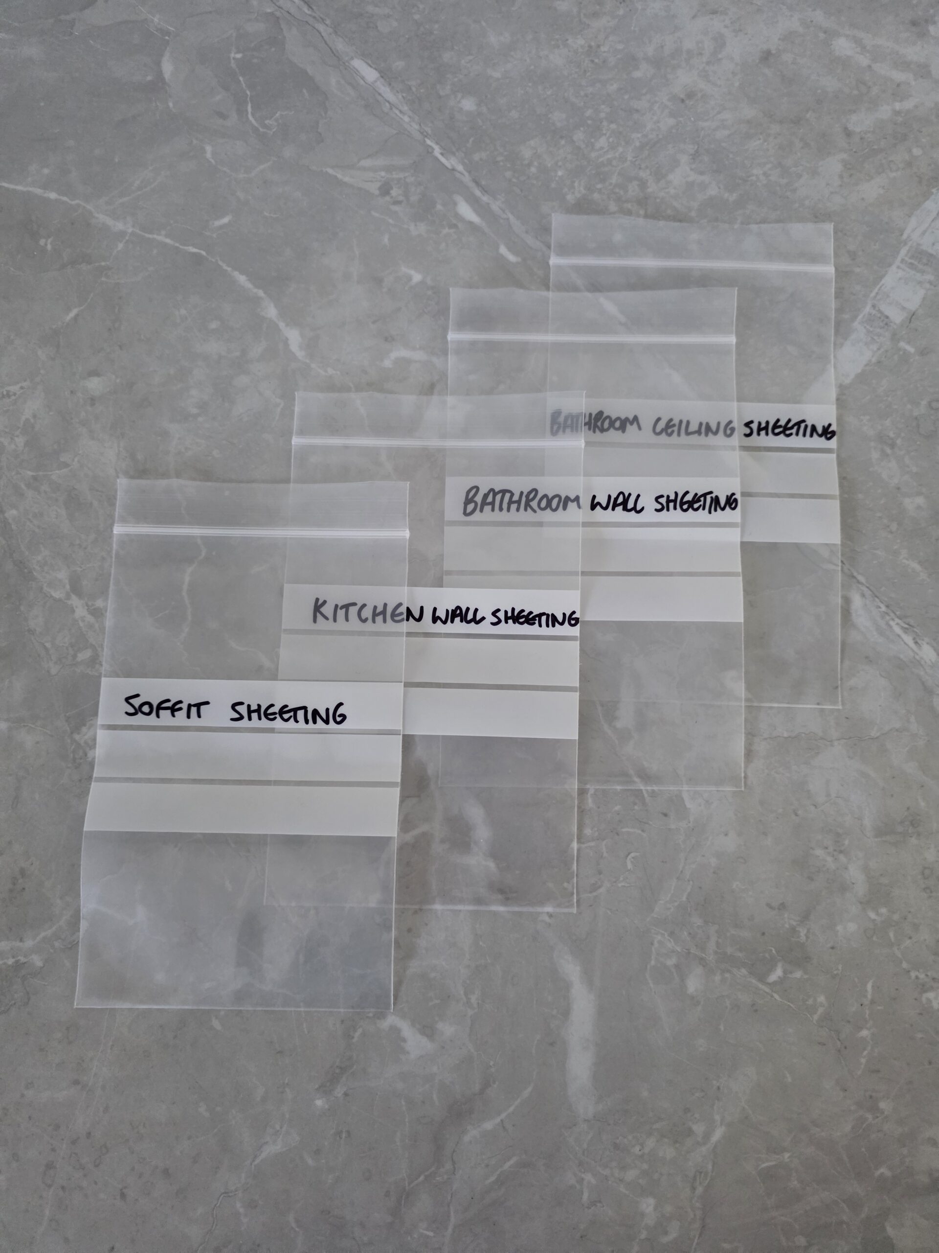 Five transparent plastic samples labelled “SOFFIT SHEETING,” “KITCHEN WALL SHEETING,” “BATHROOM WALL SHEETING,” and “BATHROOM CEILING SHEETING” are laid out on a grey marble surface in an overlapping row. Irwin Services asbestos removal Brisbane specialist working on site June 2026