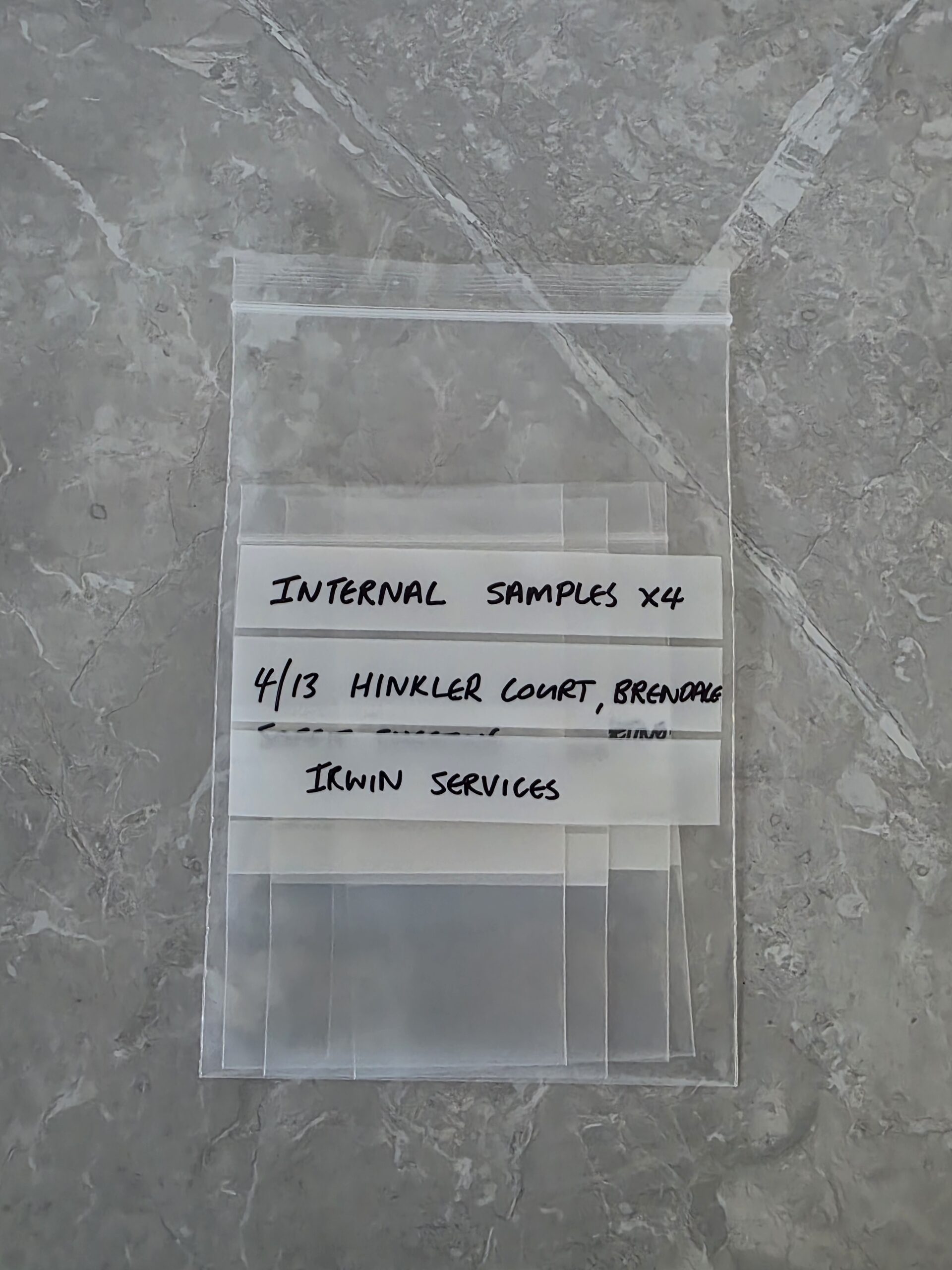 A clear resealable plastic bag labelled INTERNAL SAMPLES x4, 4/13 HINKLER COURT, BRENDALE, IKWN SERVICES sits on a grey, marbled surface. Irwin Services asbestos removal Brisbane job site June 2026